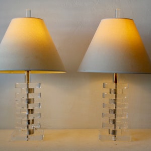 May include: Two clear acrylic table lamps with white fabric lampshades. The lamps have a stacked block design.