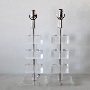 May include: A pair of clear acrylic table lamps with a stacked design. Each lamp has a silver metal base and a clear glass shade.