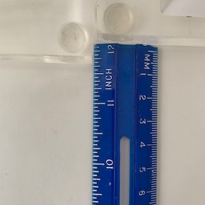 May include: A blue ruler with inch and millimetre markings. The ruler is placed against a white surface with two clear adhesive pads.