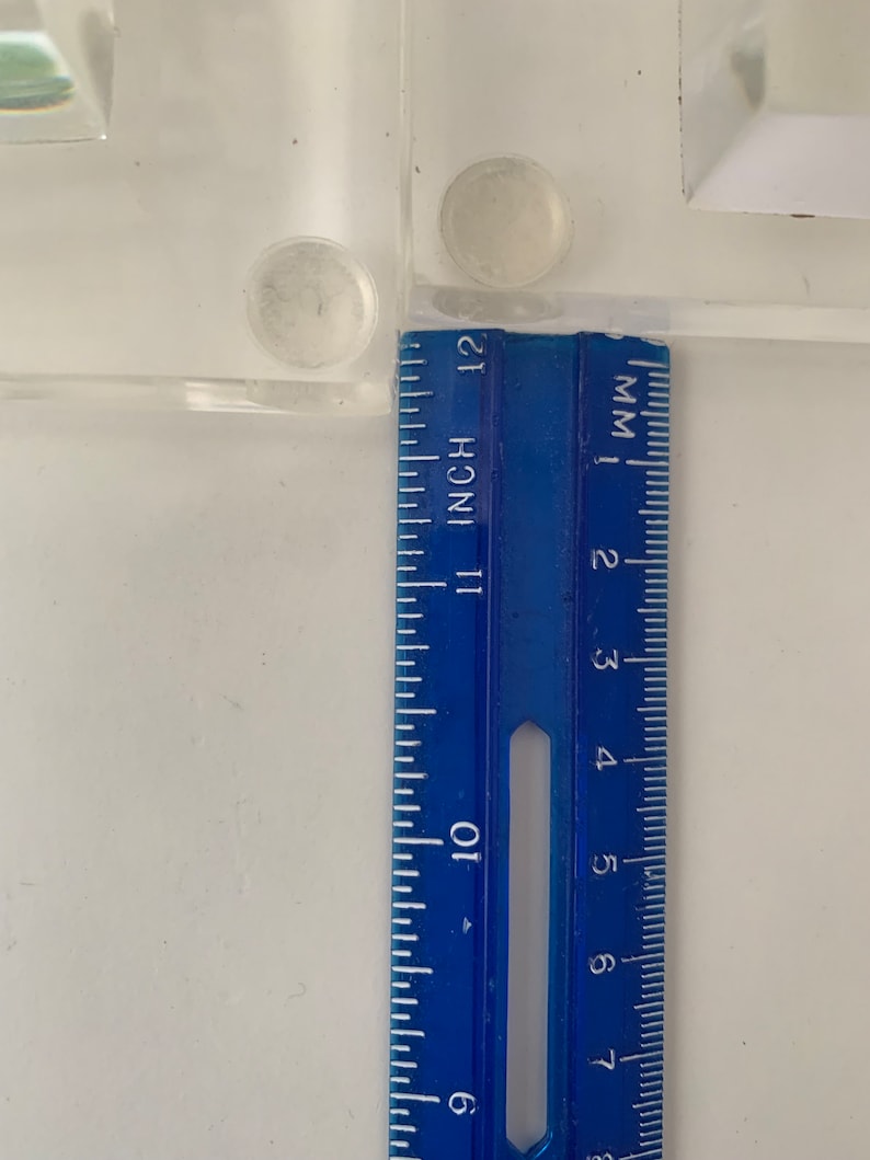 May include: A blue ruler with inch and millimetre markings. The ruler is placed against a white surface with two clear adhesive pads.
