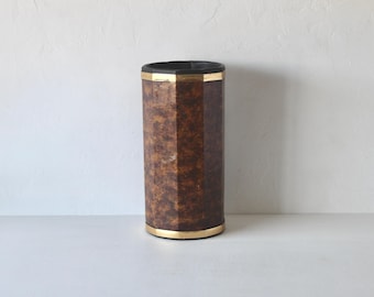 Pode incluir: Um recipiente alto e cilíndrico com um exterior castanho texturizado e faixas douradas na parte superior e inferior. A borda superior é preta. O recipiente tem um design geométrico facetado. O fundo é uma parede branca lisa.