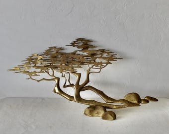Pode incluir: Uma escultura de parede de metal dourado que representa uma árvore estilizada com galhos e folhas. A árvore está dobrada e repousa sobre três pedras.