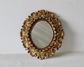 May include: Oval-shaped antique mirror with a gold-colored ornate frame. The frame features intricate scrollwork and decorative elements. The mirror's reflective surface is surrounded by a detailed, gilded border, adding to its vintage aesthetic.