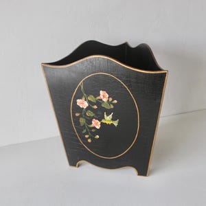 May include: Black wooden magazine holder with gold trim and a floral and hummingbird design. The oval design features pink flowers and a yellow hummingbird. The holder has a unique, curved top and is supported by small feet.