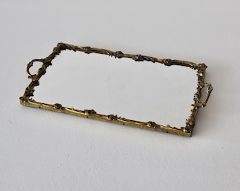 Pode incluir: Uma bandeja retangular com espelho apresentando uma moldura de metal na cor dourado antigo com detalhes florais intrincados e duas alças decorativas nos lados mais curtos. A superfície central é um painel de espelho de vidro claro e reflexivo.