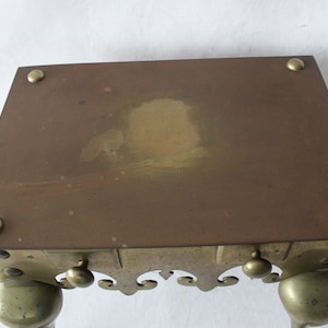 May include: A brass and copper fire screen with a decorative scrolled base and four legs. The screen has a rectangular top with a slightly worn surface.