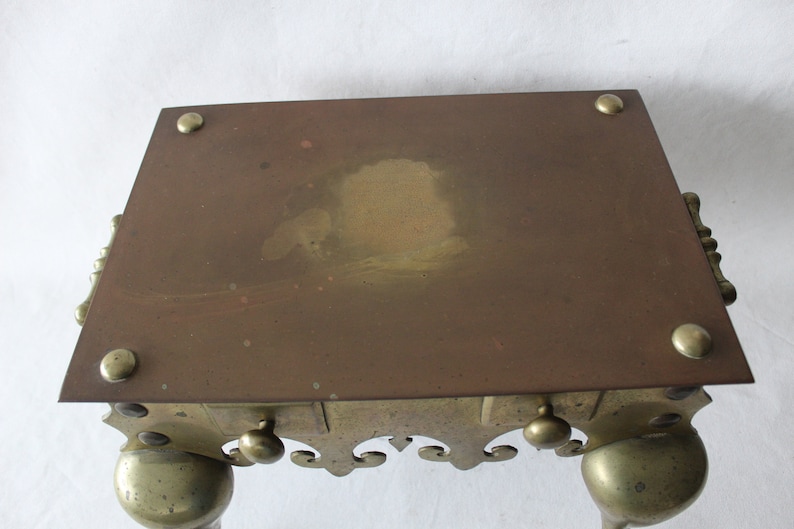 May include: A brass and copper fire screen with a decorative scrolled base and four legs. The screen has a rectangular top with a slightly worn surface.