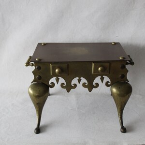 May include: A brass fireplace stand with a square top and three legs. The stand has a decorative design around the top and bottom.