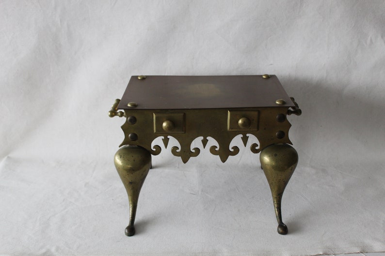 May include: A brass fireplace stand with a square top and three legs. The stand has a decorative design around the top and bottom.