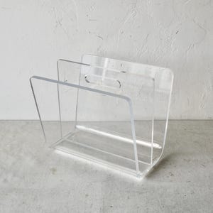 May include: A clear acrylic magazine rack with a handle. The rack is designed to hold magazines or other items upright.