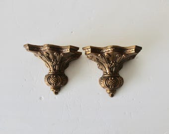 Pode incluir: Duas prateleiras de parede de metal dourado adornadas com um design decorativo. As prateleiras têm a forma de um semicírculo e uma borda festoneada.