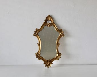 Pode incluir: Um espelho de parede com uma moldura dourada ornamentada com volutas complexas e detalhes florais. A moldura tem uma forma simétrica e alongada com uma crista decorativa no topo e um ornamento correspondente na borda inferior.