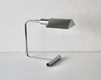 Pode incluir: Uma moderna luminária de mesa com acabamento cromado e uma tela retangular cinza. A luminária tem um braço curvo e uma base cilíndrica. A luminária tem aproximadamente 30 cm de altura.