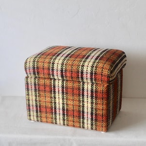 May include: A plaid storage ottoman with a brown, orange, white, and black pattern. The ottoman has a hinged lid and is made of fabric.