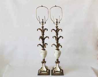 Może przedstawiać: Para białych i złotych lamp stołowych z ozdobnymi wzorami. Lampy mają białą ceramiczną podstawę z złotymi akcentami i złotą metalową ramę z wzorem liści.