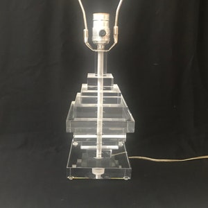 May include: A clear acrylic table lamp with a stepped base and a silver-toned lamp holder. The lamp has a rectangular base and a clear cord. The lamp is approximately 18 inches tall.