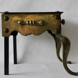 May include: A brass and black metal stand with a scalloped edge and a handle. The stand has three legs and is designed to hold a hot pot or other cooking item.