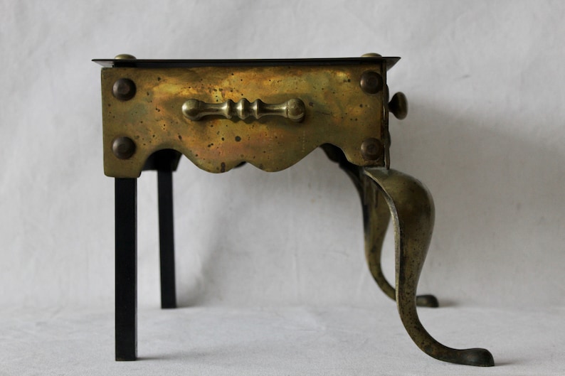 May include: A brass and black metal stand with a scalloped edge and a handle. The stand has three legs and is designed to hold a hot pot or other cooking item.