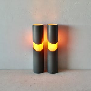 May include: Two gray cylindrical lamps with orange light emanating from the center of each lamp.