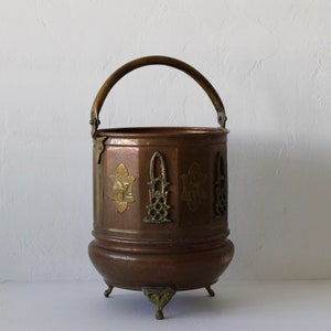 May include: A copper bucket with a brass handle and decorative accents. The bucket has a rounded bottom and sits on three feet.
