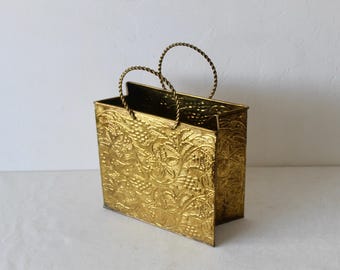 Pode incluir: Uma sacola de compras de metal dourado com um design floral em relevo. A sacola tem duas alças e é feita de metal resistente.