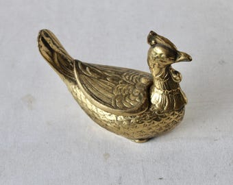 Pode incluir: Uma figurinha de pássaro de metal dourado com detalhes intrincados. O pássaro tem um pescoço comprido e uma cabeça pequena com um bico. As asas do pássaro estão abertas e suas penas da cauda são enroladas.