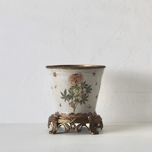 May include: A white ceramic planter with a tapered shape, featuring a painted floral design in pink and green with small red accents. The planter is set into a decorative, ornate gold-toned metal base with four curved feet and a textured rim.
