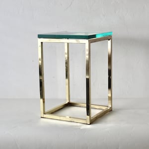 May include: A modern side table with a clear glass top and a gold-colored metal frame. The table has a square shape and a minimalist design, suitable for various interior styles. The table measures approximately 20 inches in height.