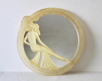 Pode incluir: Um espelho redondo com uma moldura de cor creme que apresenta um design esculpido de uma mulher com um vestido fluido. O espelho tem uma superfície refletora.