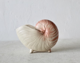 Pode incluir: Um vaso de cerâmica em forma de concha do mar com um design de espiral branco e rosa. O vaso tem três pés pequenos e é perfeito para exibir plantas pequenas.