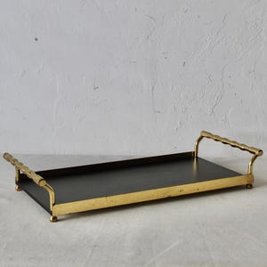 Può includere: Vassoio rettangolare con interno nero e cornice in metallo color oro. Il vassoio ha due manici con un design strutturato. Il vassoio è lungo circa 40 cm e largo 25 cm.