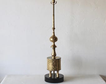 Pode incluir: Um candeeiro de mesa alto de ouro com um design ornamentado. O candeeiro tem uma base redonda com três pernas e um abajur cilíndrico. O abajur é decorado com um padrão floral. O candeeiro está sobre uma toalha de mesa branca.