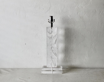 Puede incluir: Una lámpara de mesa de acrílico transparente con una base rectangular y un cuerpo alto y rectangular. El cuerpo presenta líneas verticales grabadas y diseños curvos. La lámpara tiene un casquillo plateado.