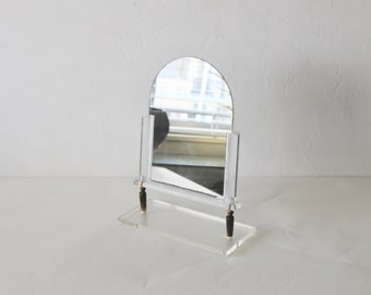 May include: A clear acrylic tabletop mirror with an arched top and a small shelf. The mirror is on a white surface.