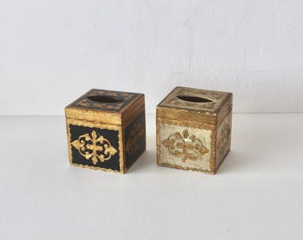 Pode incluir: Duas caixas quadradas para lenços com detalhes ornamentais em folha de ouro. Uma caixa tem uma base preta mate, enquanto a outra tem uma base prateada metálica. Ambas apresentam uma abertura oval central e padrões dourados em relevo.