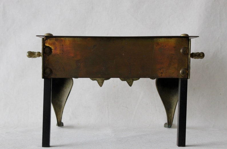May include: A vintage brass and black metal fireplace stand with a rectangular top and four legs. The stand has a decorative design on the front and back.