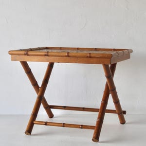 Op de afbeelding: Een rechthoekige klaptafel gemaakt van bruin hout in bamboe-stijl. De tafel heeft een vlak bovenblad met een opstaande rand en een onderstel bestaande uit gekruiste poten die verbonden zijn door horizontale steunbalken.