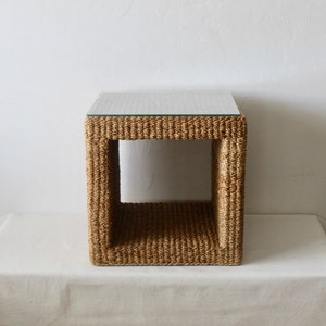 May include: A square end table made of woven natural fiber with a glass top. The table has a simple, open design with a woven base and a clear glass top.