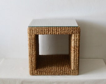 Pode incluir: Uma mesa auxiliar quadrada feita de fibra natural tecida com tampo de vidro. A mesa tem um design simples e aberto com uma base tecida e um tampo de vidro transparente.