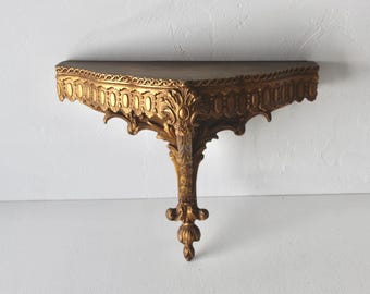 Pode incluir: Prateleira de parede dourada ornamentada com um tampo castanho escuro. A prateleira apresenta entalhes intrincados e uma única perna decorativa. Montada numa parede branca, mostra o seu design detalhado e estética vintage.