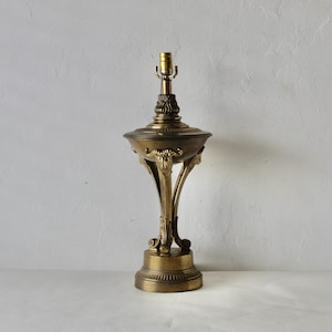 May include: A vintage brass table lamp with an ornate design. The lamp features a detailed base, a decorative bowl-shaped center, and a textured top with a light bulb socket. The lamp is a warm brass color and stands against a neutral background.
