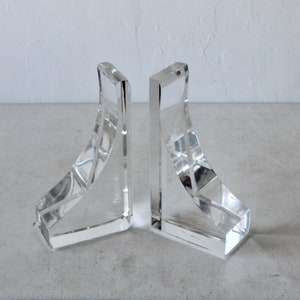 May include: A pair of clear glass bookends with a geometric design. The bookends are angled and have a curved shape.