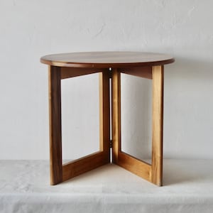 Può includere: Un tavolino rotondo in legno, con un piano marrone scuro e gambe marroni chiare. Le gambe sono rettangolari e formano un design geometrico aperto. Il tavolo misura circa 50 cm di diametro e 61 cm di altezza.