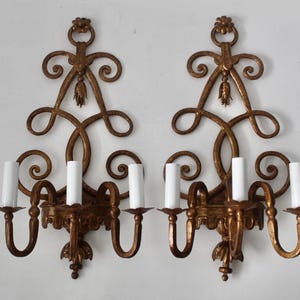 Puede incluir: Un par de apliques de pared de metal dorado con volutas intrincadas, detalles florales en la parte superior e inferior, y tres cubiertas de luz blancas estilo vela en cada lámpara. El metal tiene un acabado texturizado con reflejos oscuros.
