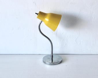 Pode incluir: Uma lâmpada de mesa amarela vintage com um pescoço de ganso flexível prateado e uma base redonda prateada. A sombra da lâmpada é amarelo brilhante e a base é prateada brilhante. A lâmpada é ajustável.