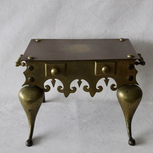 May include: A brass fireplace stand with a square top and three legs. The stand has a decorative design around the edge and a raised center on the top.