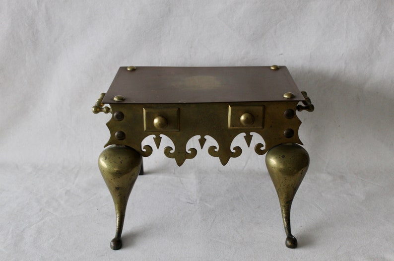 May include: A brass fireplace stand with a square top and three legs. The stand has a decorative design around the edge and a raised center on the top.