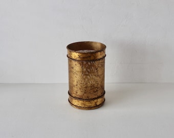 Pode incluir: Um recipiente cilíndrico de metal em tom dourado com um acabamento texturizado e desgastado. O objeto apresenta duas faixas horizontais com detalhes de corda trançada nas bordas superior e inferior, conferindo-lhe uma aparência estruturada.