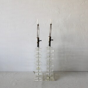 May include: A pair of clear acrylic table lamps with a stacked block design. The lamps have a silver finish and a simple design.