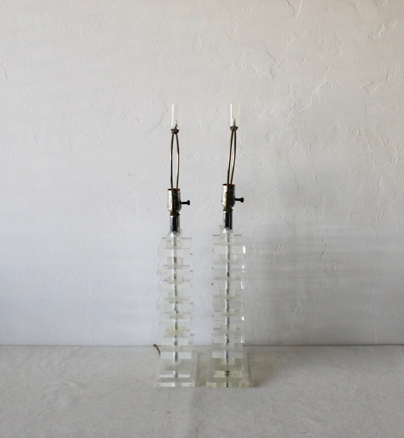 May include: A pair of clear acrylic table lamps with a stacked block design. The lamps have a silver finish and a simple design.